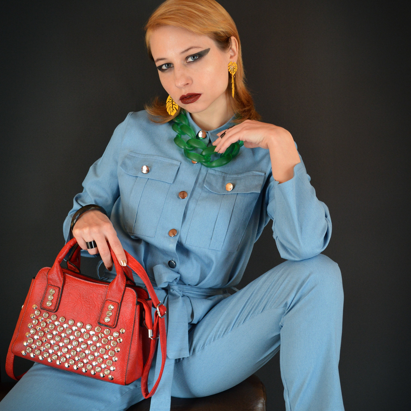 Woman in a blue outfit holding a red studded handbag against a dark background